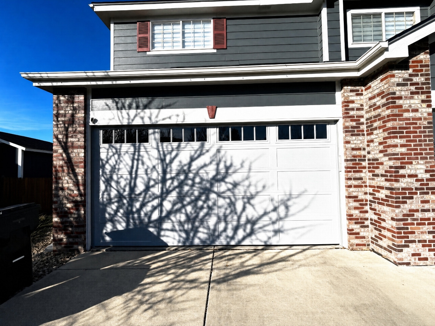 Same garage door photographed in harsh direct sunlight with heavy tree branch shadows striping the door
